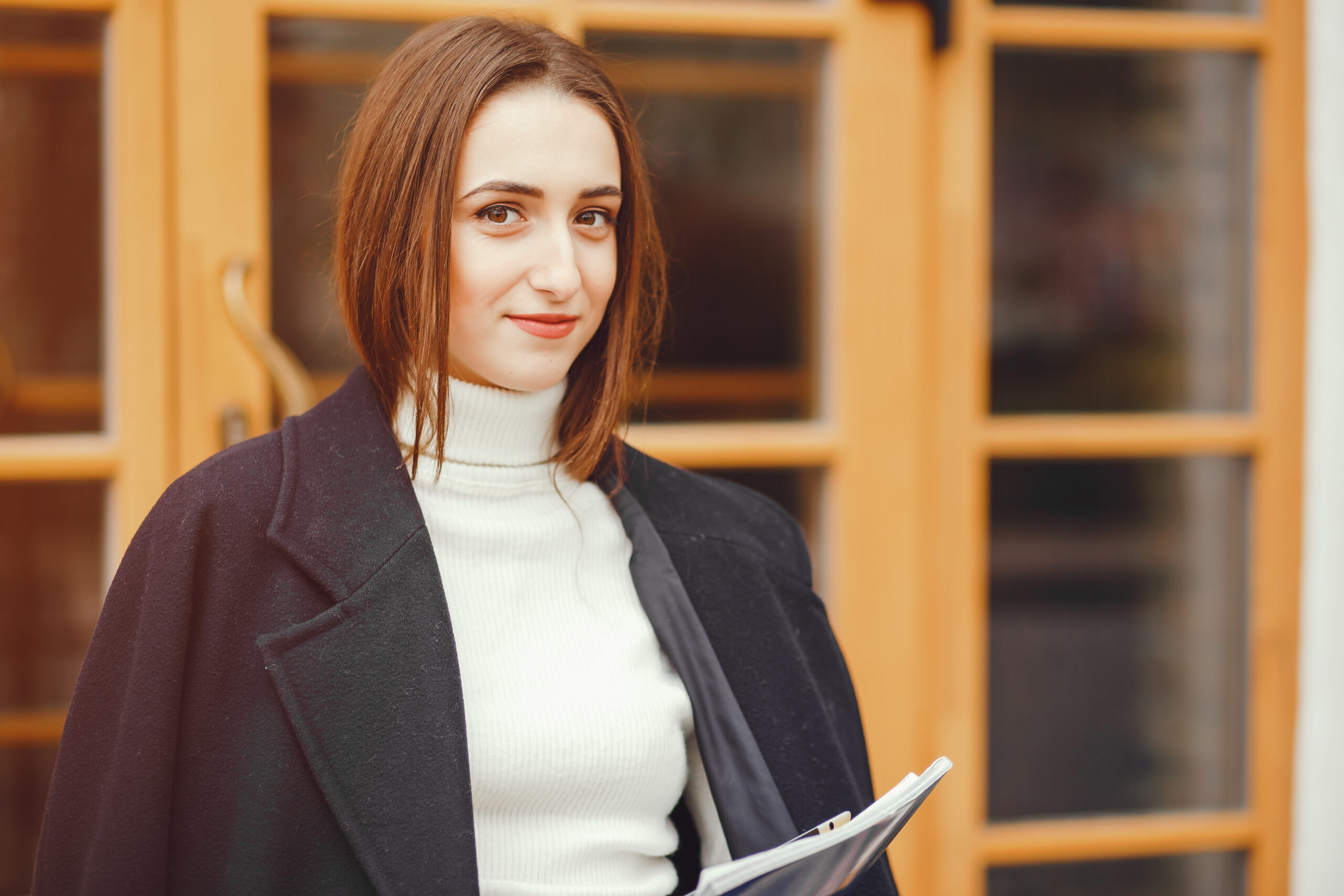 Beautiful girl in a black coat. Cute lady near building. Woman at the door